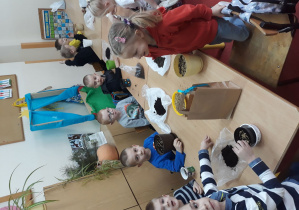 Na zdjęciu widać grupę dzieci znajdującą się w sali przedszkolnej, które siedzą przy stolikach. Każde z nich ma przed sobą donicę z ziemią oraz owies w miseczkach plastikowych. Dzieci będą siały owies w donicach.
